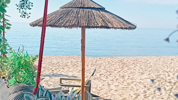 Escena de playa con una sombrilla de paja, una silla de madera y una terraza de madera.