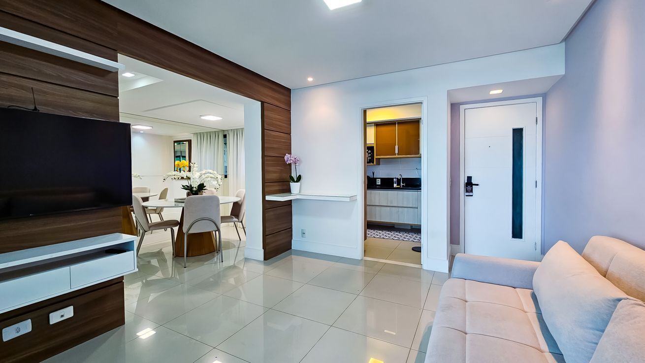 Una sala de estar moderna con televisor de pantalla plana, un sofá, un comedor con mesa y sillas, y una cocina visible a través de una puerta abierta.