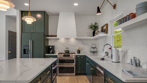 Une cuisine moderne avec des meubles de couleur verte, un plan de travail blanc, des appareils en acier inoxydable et des luminaires suspendus.