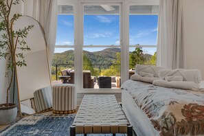 Ein Schlafzimmer mit großem Fenster, einem Bett, einem Nachttisch, einem Sessel, einem kleinen Tisch und einer Topfpflanze.