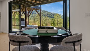 Une table ronde recouverte d’un drap de feutre vert, entourée de quatre chaises rembourrées, dans une pièce dotée de grandes fenêtres donnant sur une colline boisée.