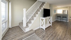 Un amplio salón con una escalera de madera, un televisor de pantalla plana sobre una mesa consola blanca y una cocina con electrodomésticos de acero inoxidable.