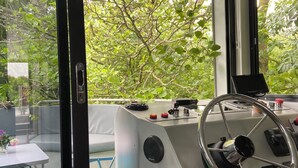 A control station with a steering wheel, a tablet, and various controls, overlooking a green outdoor area with trees and a pool.