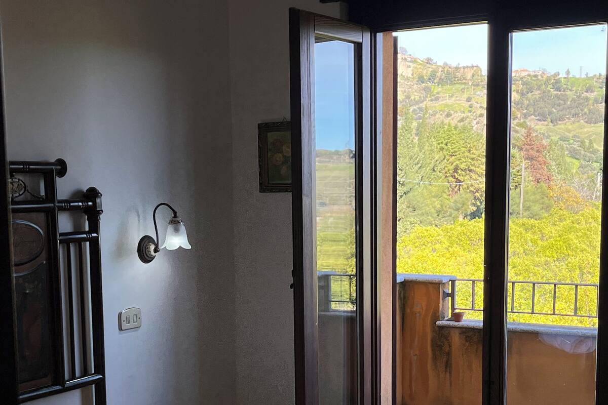 Ein Hotelzimmer mit Bett, Nachttisch, Wandlampe und Blick auf einen Balkon mit Grünpflanzen.