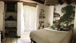 Un dormitorio con cama, una silla, ventana con cortinas, pared con caligrafía y un cuadro de árboles.