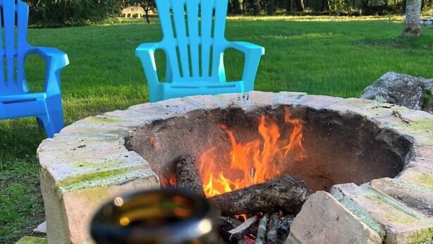 Un hogar con leña ardiendo, rodeado de muros de piedra, y dos sillas Adirondack azules en un espacio al aire libre cubierto de césped.
