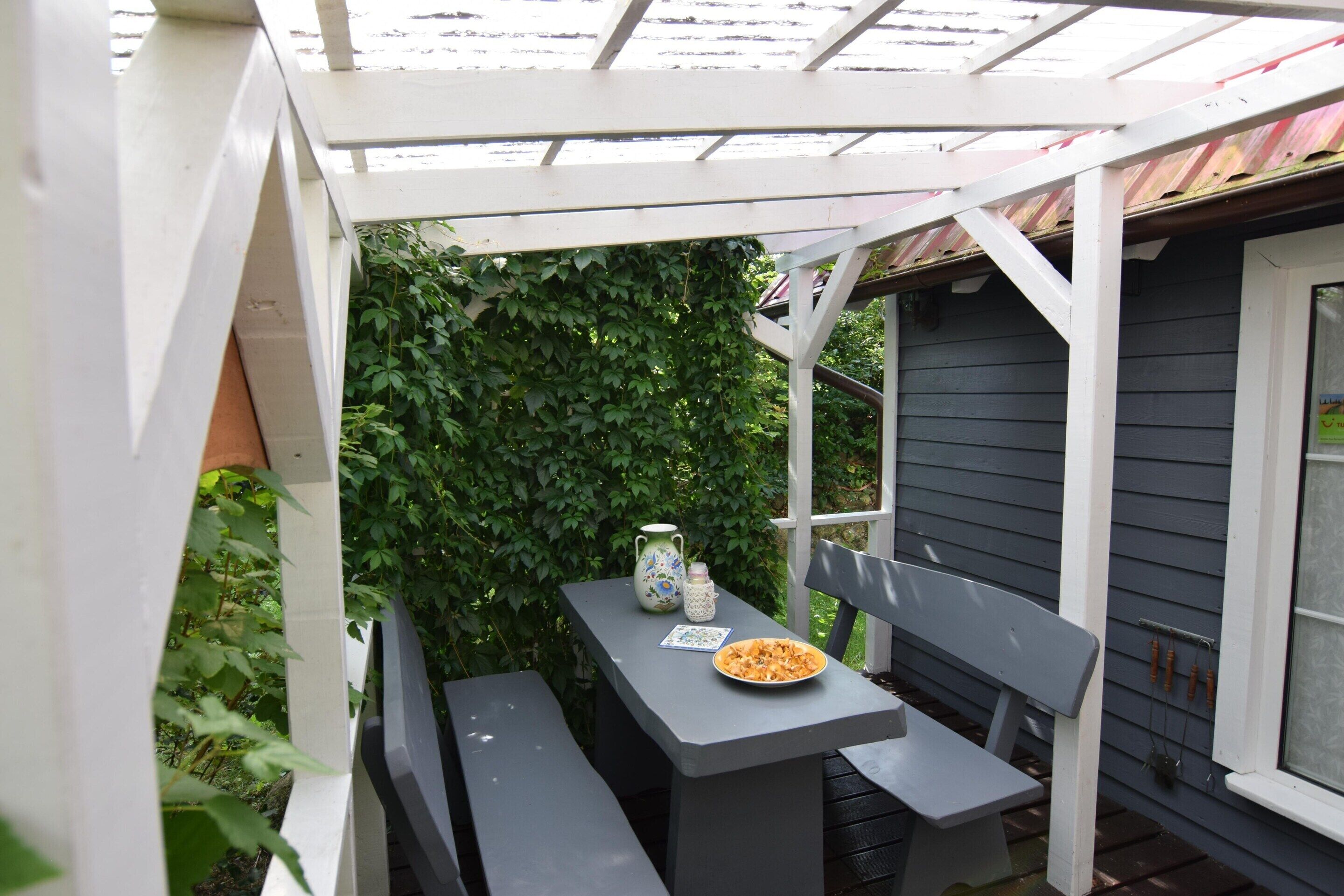 A covered patio with a table and bench, surrounded by greenery.