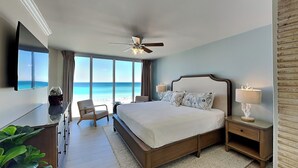 Habitación de hotel con una cama grande, ventilador de techo, televisor, escritorio con planta y vista a la playa.