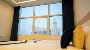 Ein Hotelzimmer mit großem Fenster, einem Bett mit Kissen und Blick auf eine Stadtlandschaft, darunter ein hoher Turm.