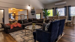 Ein geräumiges Wohnzimmer mit einem dunkelblauen Sofa, einem Glastisch und Holzboden. Ein Deckenventilator, ein Essbereich im Hintergrund und ein großes Fenster mit Vorhängen sind ebenfalls vorhanden.