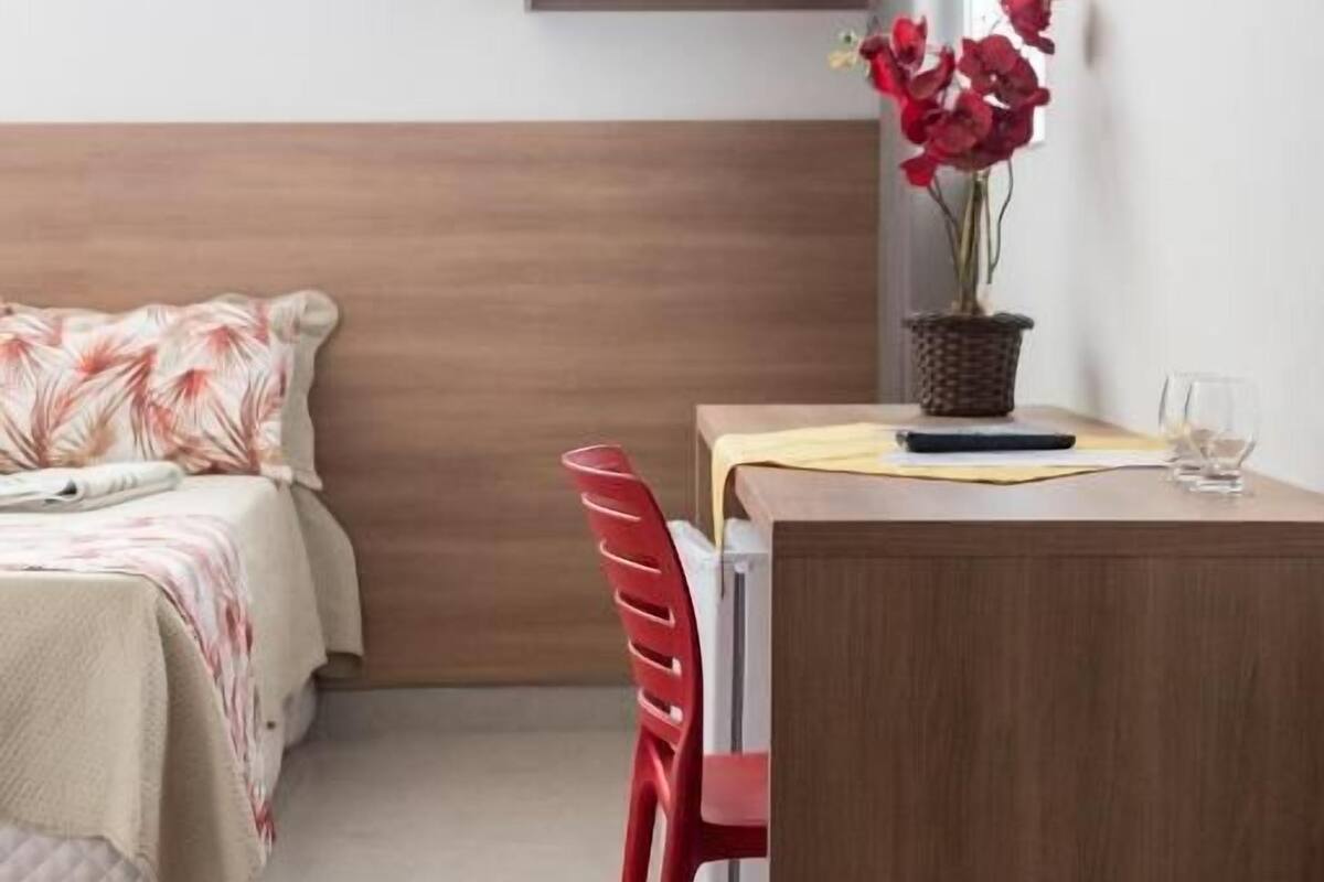 A bedroom with a bed, a desk, a chair, a bedside table, a vase with flowers, and a window.