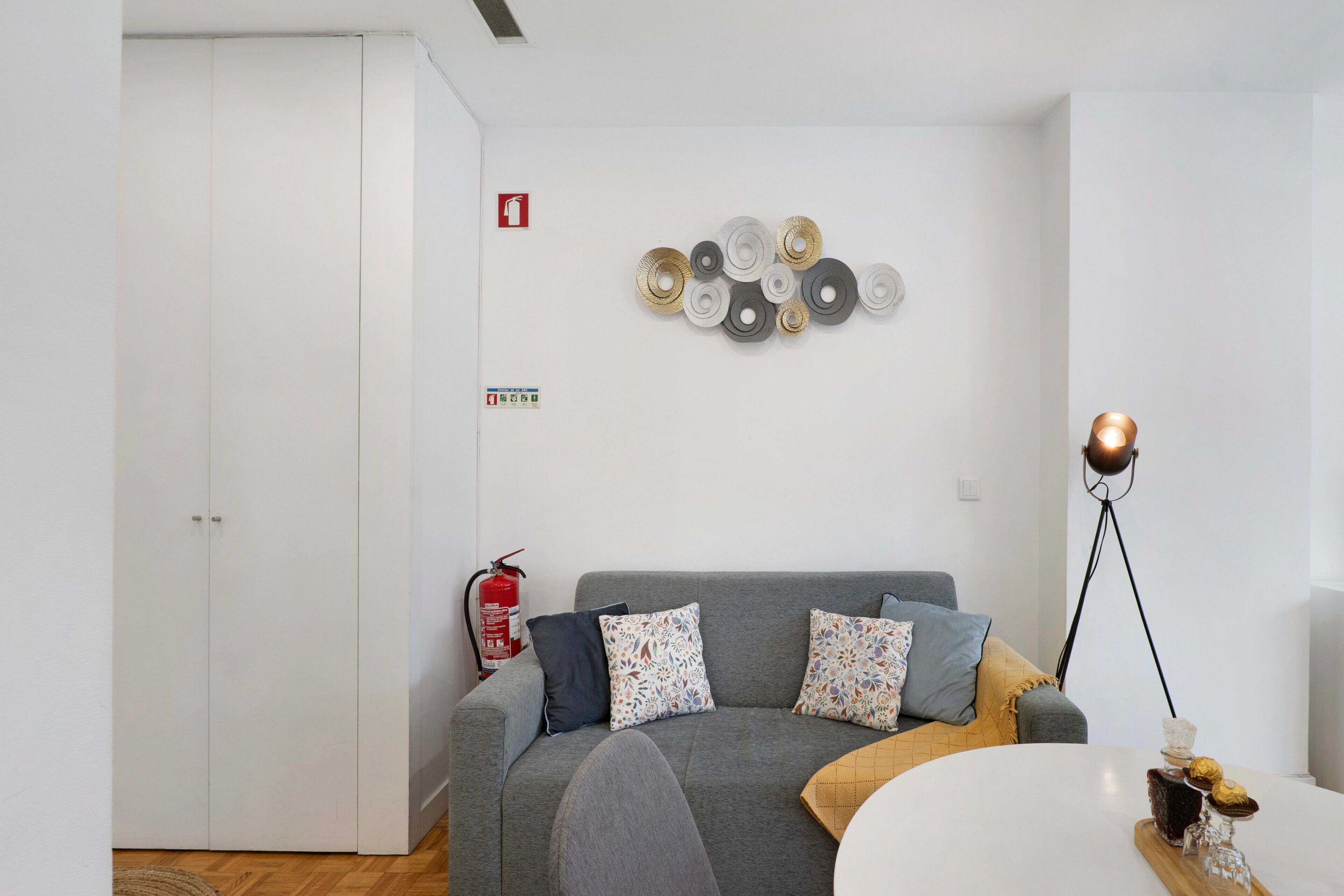 Ein modernes Wohnzimmer mit einem grauen Sofa, gemusterten Kissen, einem weißen runden Tisch, einer Wanddekoration und einer Stehlampe.