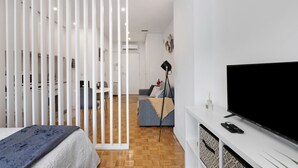 Ein modernes Zimmer mit Bett, Fernseher, Sofa und einer Trennwand mit vertikalen Lamellen.