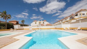 Ein Swimmingpool mit klarem, blauem Wasser, umgeben von Gebäuden in Weiß und Gelb mit Balkonen, Palmen und blauem Himmel.