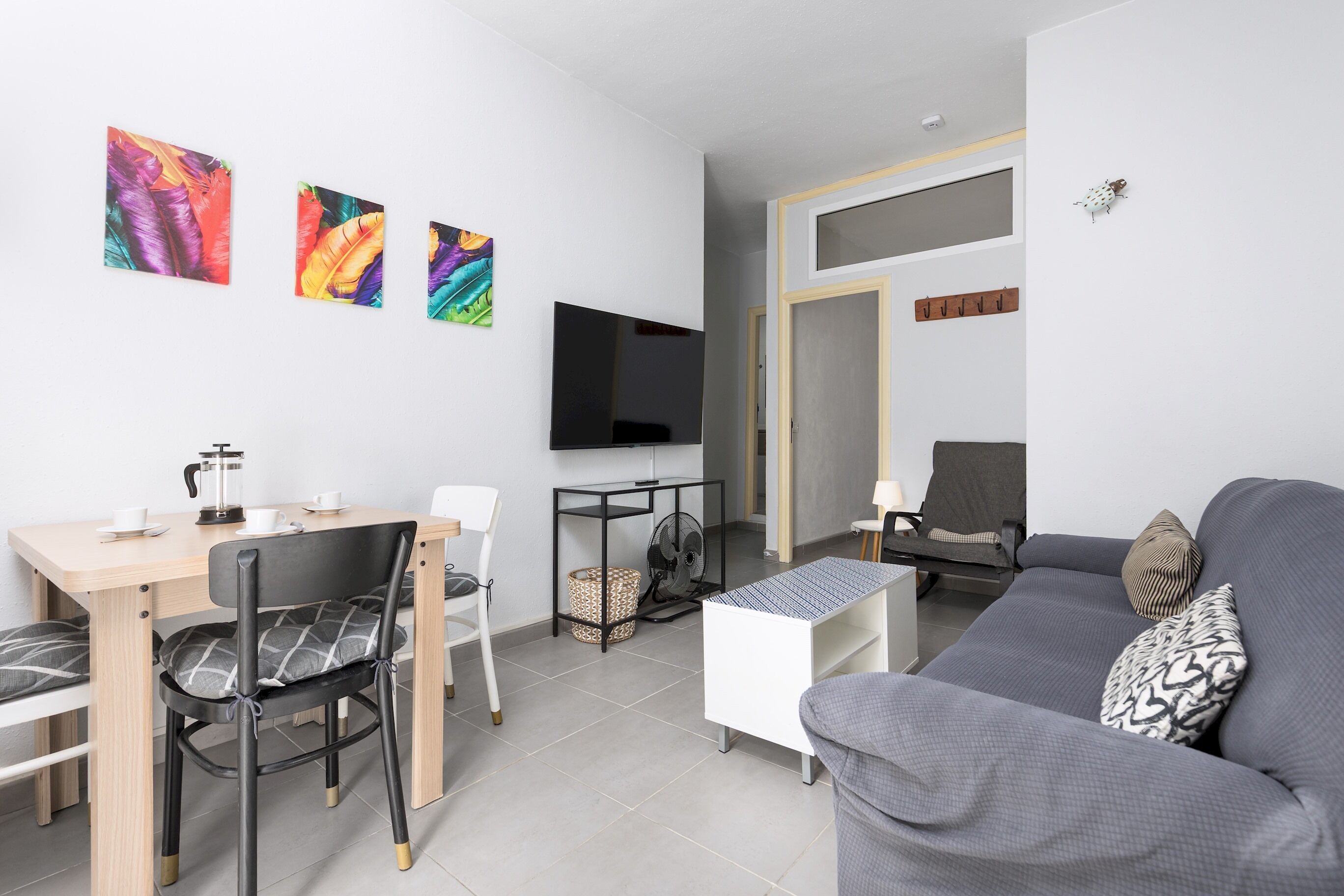 A modern living room with a grey sofa, a dining table with chairs, a coffee table, a TV mounted on the wall, and colorful artwork.