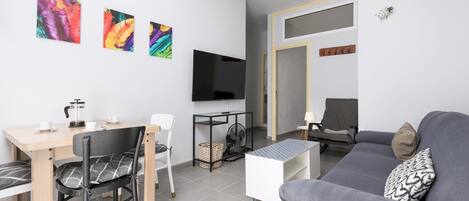 A modern living room with a grey sofa, a dining table with chairs, a coffee table, a TV mounted on the wall, and colorful artwork.