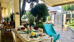 Un área de comedor al aire libre con una mesa dispuesta para dos personas, rodeada de plantas en macetas y un espacio de estar cubierto.