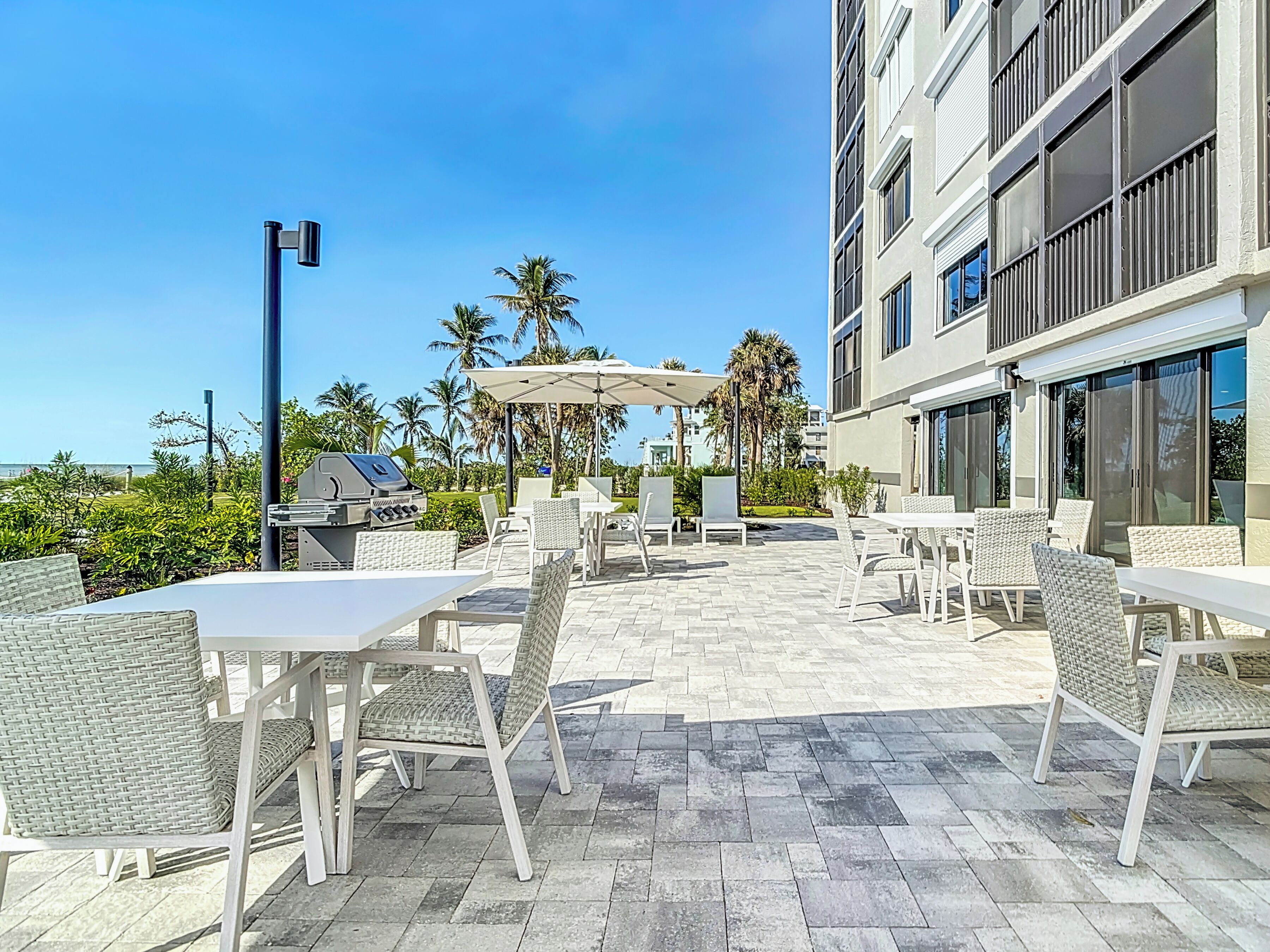An outdoor patio area with wicker furniture, a barbecue grill, and a view of palm trees and a building in the background.