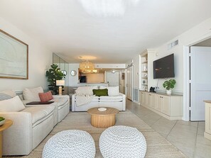 A modern living room with a sofa, coffee table, and ottomans, connected to a kitchen and dining area. - Beachfront Getaway at Caper Beach Club #803 | Private Chiller/Heated Pool (Fort Myers Beach)