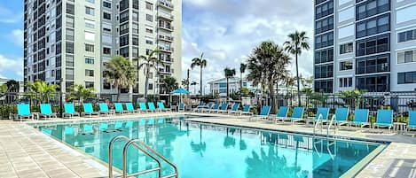 A swimming pool with blue lounge chairs and a metal ladder, surrounded by tall buildings and palm trees.