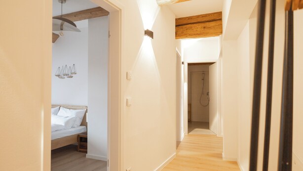Un pasillo con piso de madera, cama con ropa de cama blanca y una lámpara colgante.
