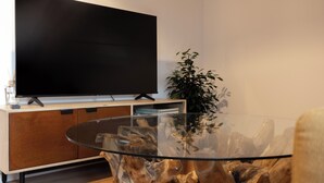 Una sala de estar moderna con un televisor de pantalla plana grande montado en un mueble de madera, una mesa de centro de vidrio con un diseño de madera único, y una planta en maceta.