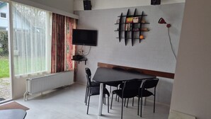Ein Zimmer mit Esstisch, Stühlen, Fernseher, Heizkörper und einem fenster mit Vorhängen.