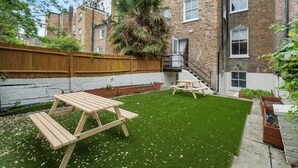A backyard with artificial grass, wooden picnic tables, a wooden fence, and a brick building in the background. - Argyl | Cambridge Gardens: Apt. B (Greater London)