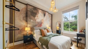 A bedroom with a large abstract painting, a bed with multiple pillows, a desk with a chair, and a window with a view of greenery. - Argyl | Cambridge Gardens: Apt. B (Greater London)
