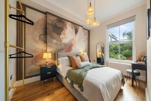 A bedroom with a large abstract painting, a bed with multiple pillows, a desk with a chair, and a window with a view of greenery. - Argyl | Cambridge Gardens: Apt. B (Greater London)