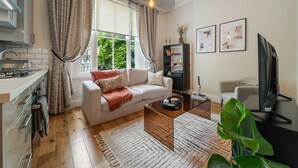 A modern living room with a sofa, coffee table, and TV. There's a kitchen area with a stove and cabinets. - Argyl | Cambridge Gardens: Apt. B (Greater London)