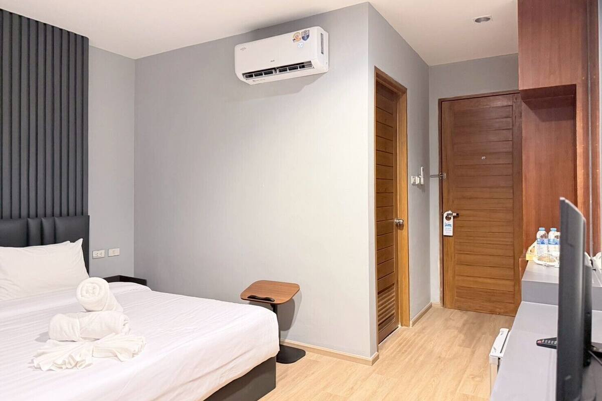 A hotel room with a bed, a small bedside table, an air conditioning unit, a wooden door, and a television.