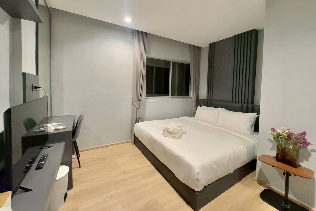 A modern hotel room with a bed, a desk with a chair, a small table with a vase of flowers, and a window with curtains.