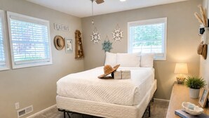 A bedroom with a bed, a ceiling fan, two windows with blinds, a bedside table with a lamp, and wall decorations. - Pet-Friendly Cozy Cape Escape Near Beach (Lower Township)