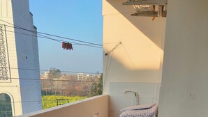 A balcony with two chairs, a table with a floral tablecloth, and a view of buildings and greenery in the background. - Twin Nest 2BHK | AC |Gated | Near Chandigarh Univ. (Mohali)