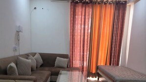 A living room with a sofa, ottomans, a coffee table, a ceiling fan, and a bed with patterned bedding. - Twin Nest 2BHK | AC |Gated | Near Chandigarh Univ. (Mohali)