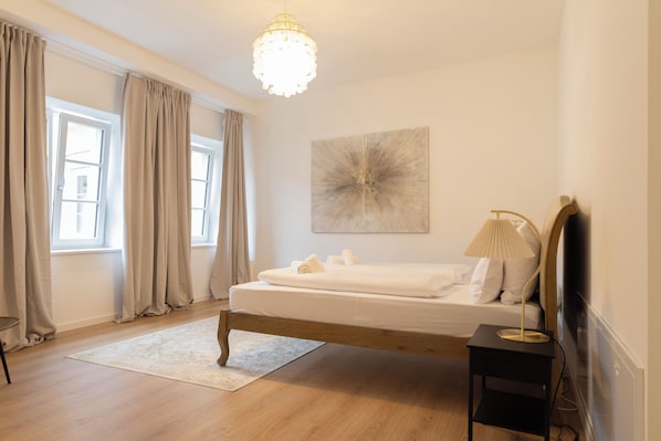 A bedroom with a wooden bed, a bedside lamp, a large window with curtains, and a wall-mounted artwork.