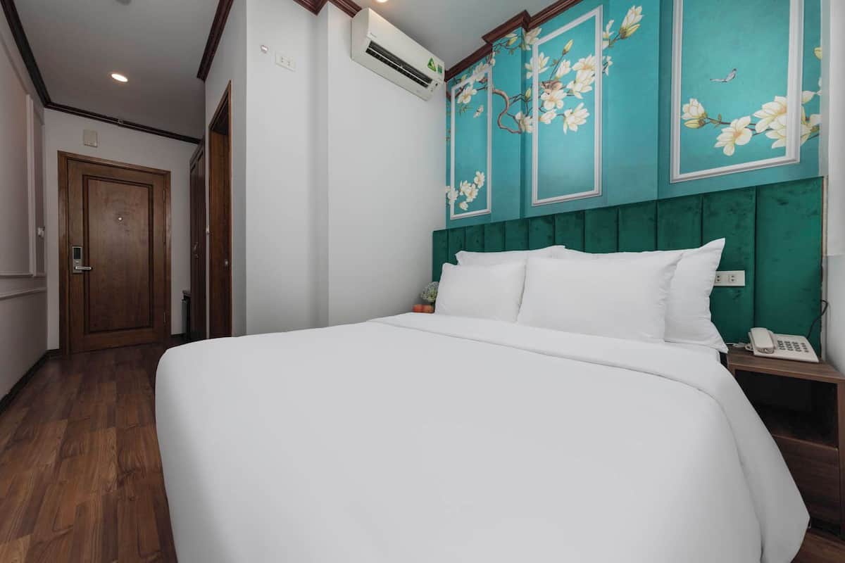 A hotel room with a large bed, a wooden floor, a teal-colored headboard with floral designs, a white wall, and a wooden door.