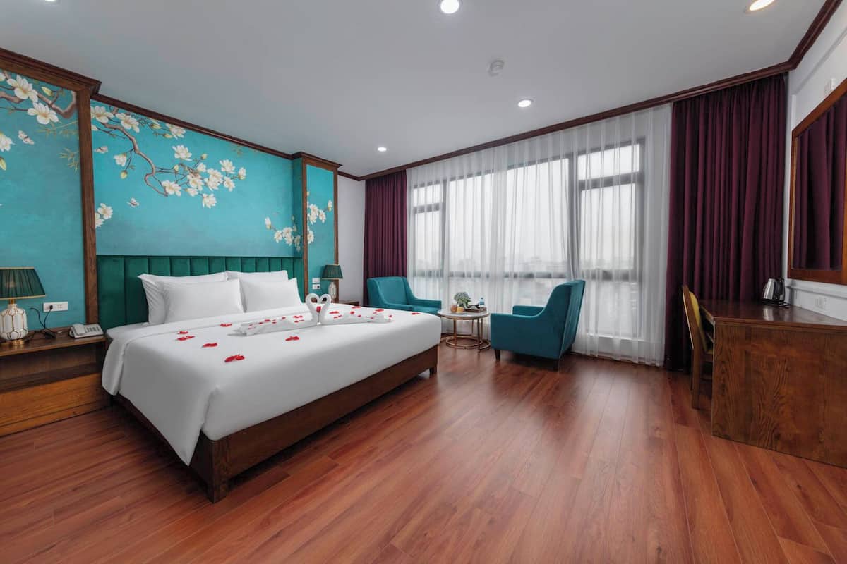 A hotel room with a large bed, a desk, two chairs, a small table, and a view of the city through large windows.