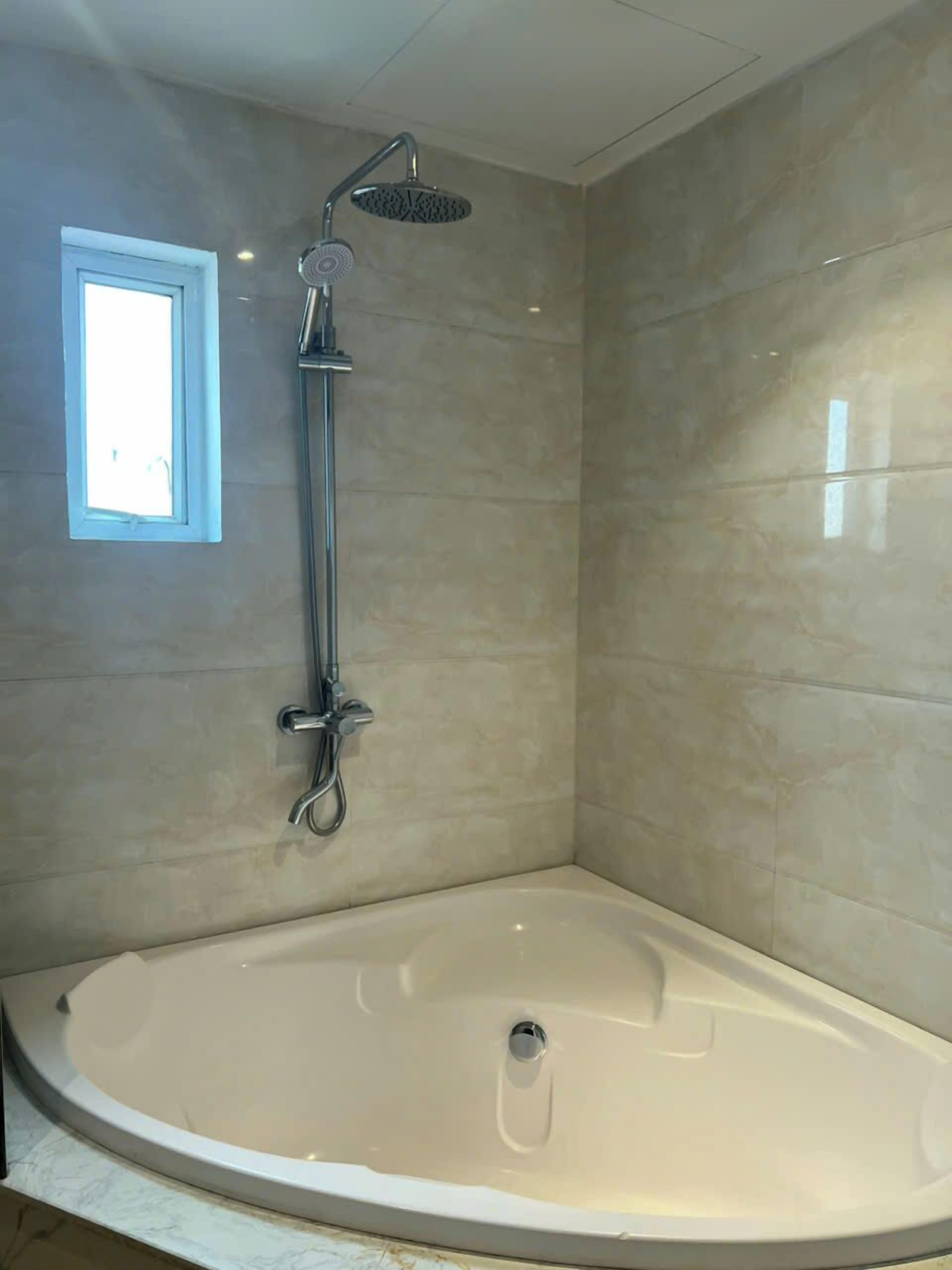A corner shower with a handheld showerhead, a small window, and a bathtub.