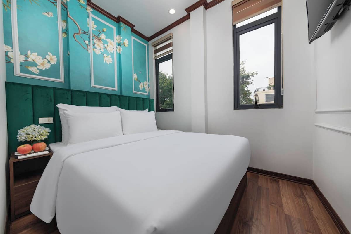 A neatly made bed in a room with teal walls, a window, and a small table with apples and flowers.