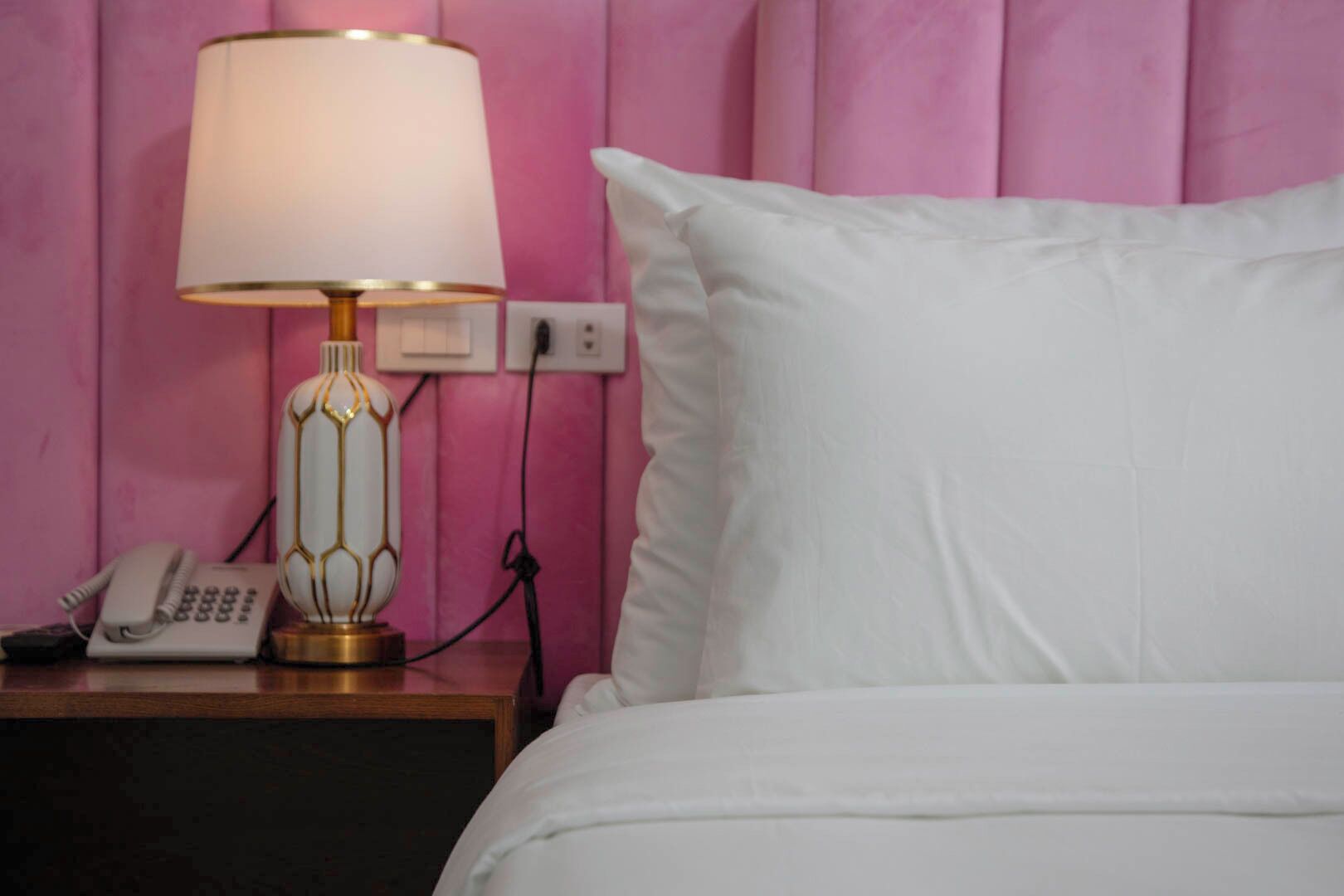 A hotel room with a white bed, a bedside lamp, a telephone, and a power outlet on the nightstand.