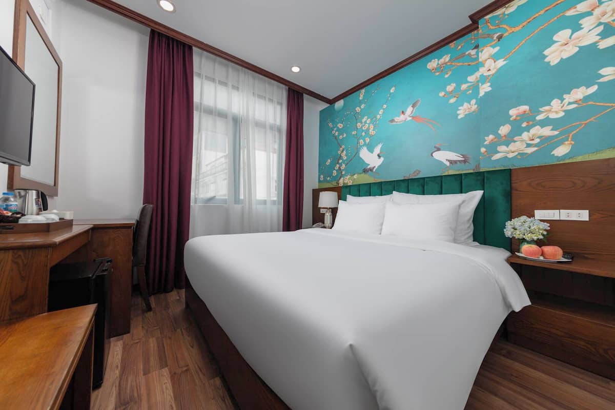 A hotel room with a large bed, a desk with a chair, a TV, and a wall with a floral mural.