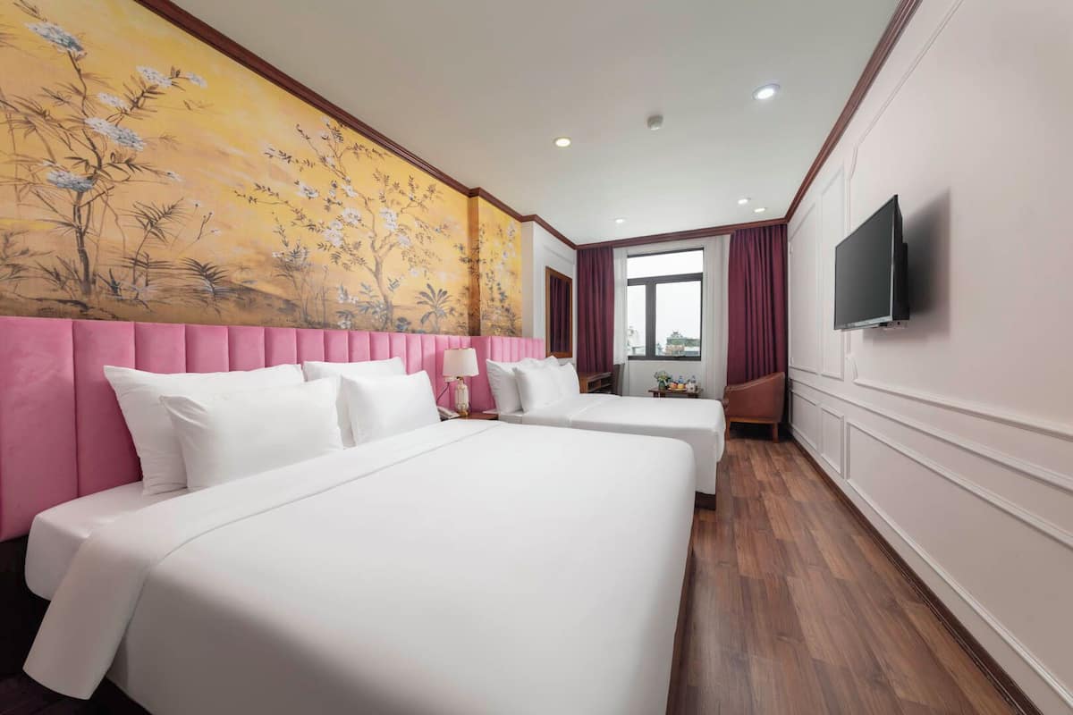 A hotel room with two beds, a pink headboard, a large painting, a TV, and a window with curtains.