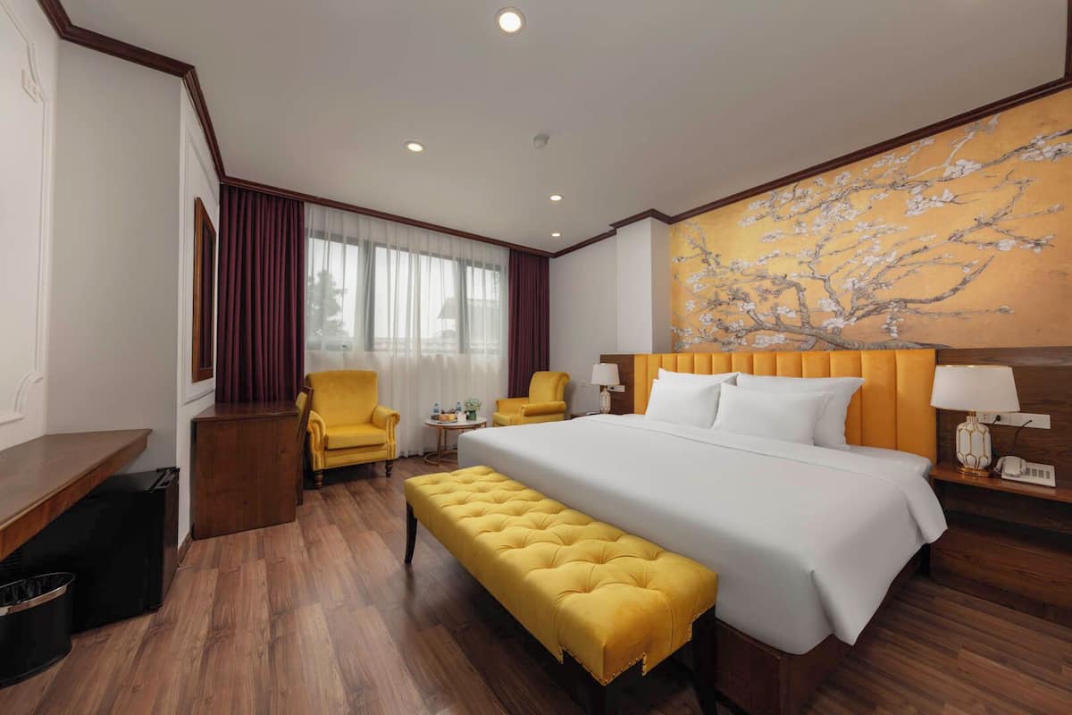A hotel room with a large bed, two armchairs, a small table, a bench, and a painting on the wall.
