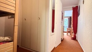 Un couloir étroit, avec un lit superposé en bois sur le côté gauche, une armoire blanche et une porte donnant sur une pièce où se trouve un rideau rouge.