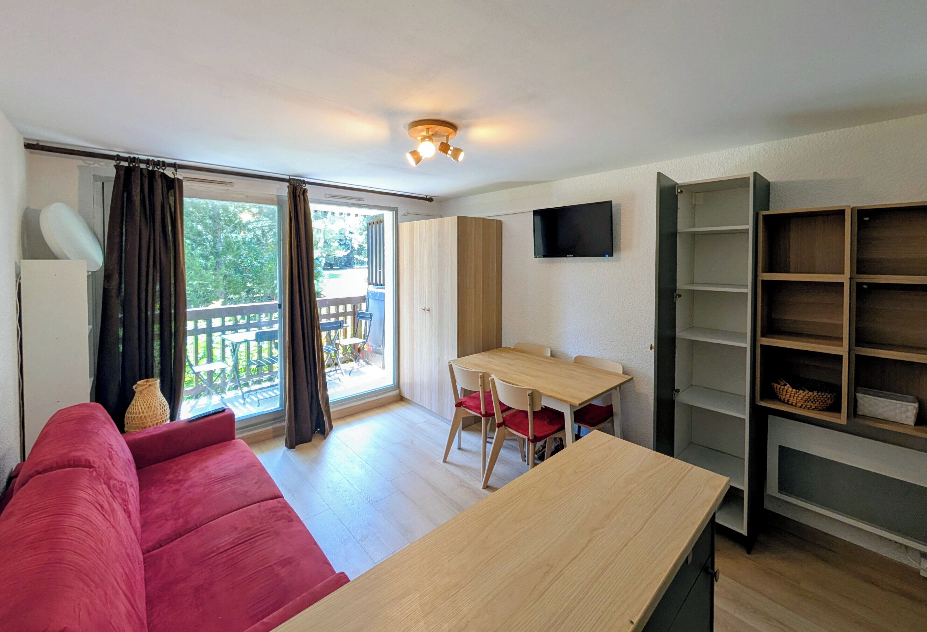 Una habitación con un sofá rojo, una mesa de madera, un televisor montado en la pared y un balcón con vistas a zonas verdes.