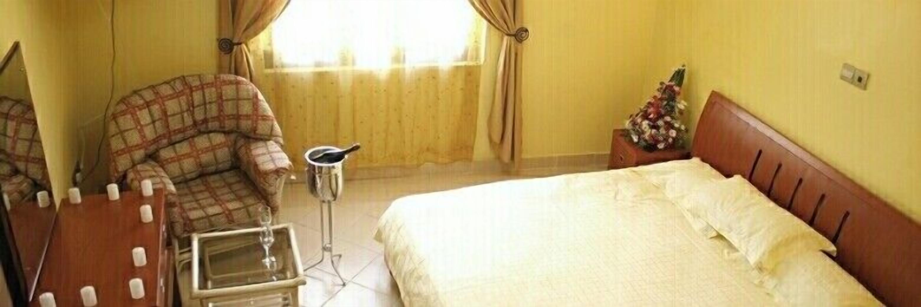 A hotel room with a bed, a chair, a small table with a glass, and a window with curtains.