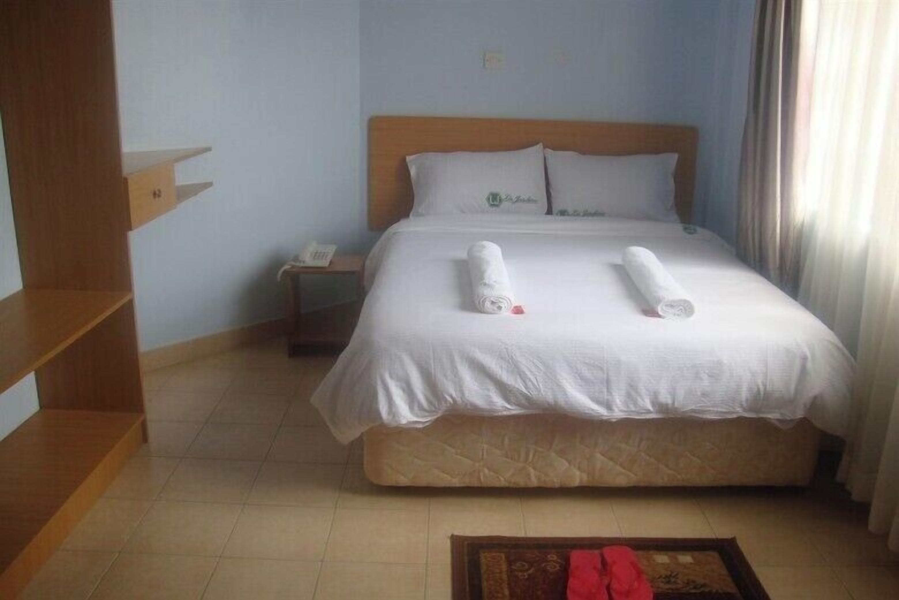 Una cama bien tendida con sábanas blancas y dos toallas enrolladas, un cabecero de madera, una mesita de noche pequeña con un teléfono, y una mesita de noche.