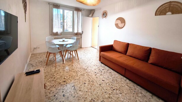 Ein Wohnzimmer mit rundem Tisch, weißen Stühlen, einem braunen Sofa, einem Fernseher und einer Deckenleuchte.